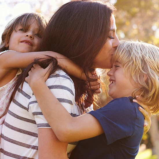 A woman hugs two smiling children outdoors in Katy, TX, kissing one on the forehead. The children’s arms are wrapped around her as sunlight shines in the background, creating a warm, joyful atmosphere.
