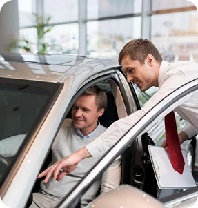 A car salesman with a red tie shows features of a car to a smiling man in the driver’s seat inside a Katy TX showroom, highlighting how Casillas Insurance services add value. The salesman holds a clipboard and points at the dashboard.