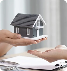 Two hands are shown: one holding a small model house and the other reaching out beneath it, suggesting buying, selling, or transferring property. A clipboard with papers is visible in the background, representing Casillas Insurance services in Katy TX.
