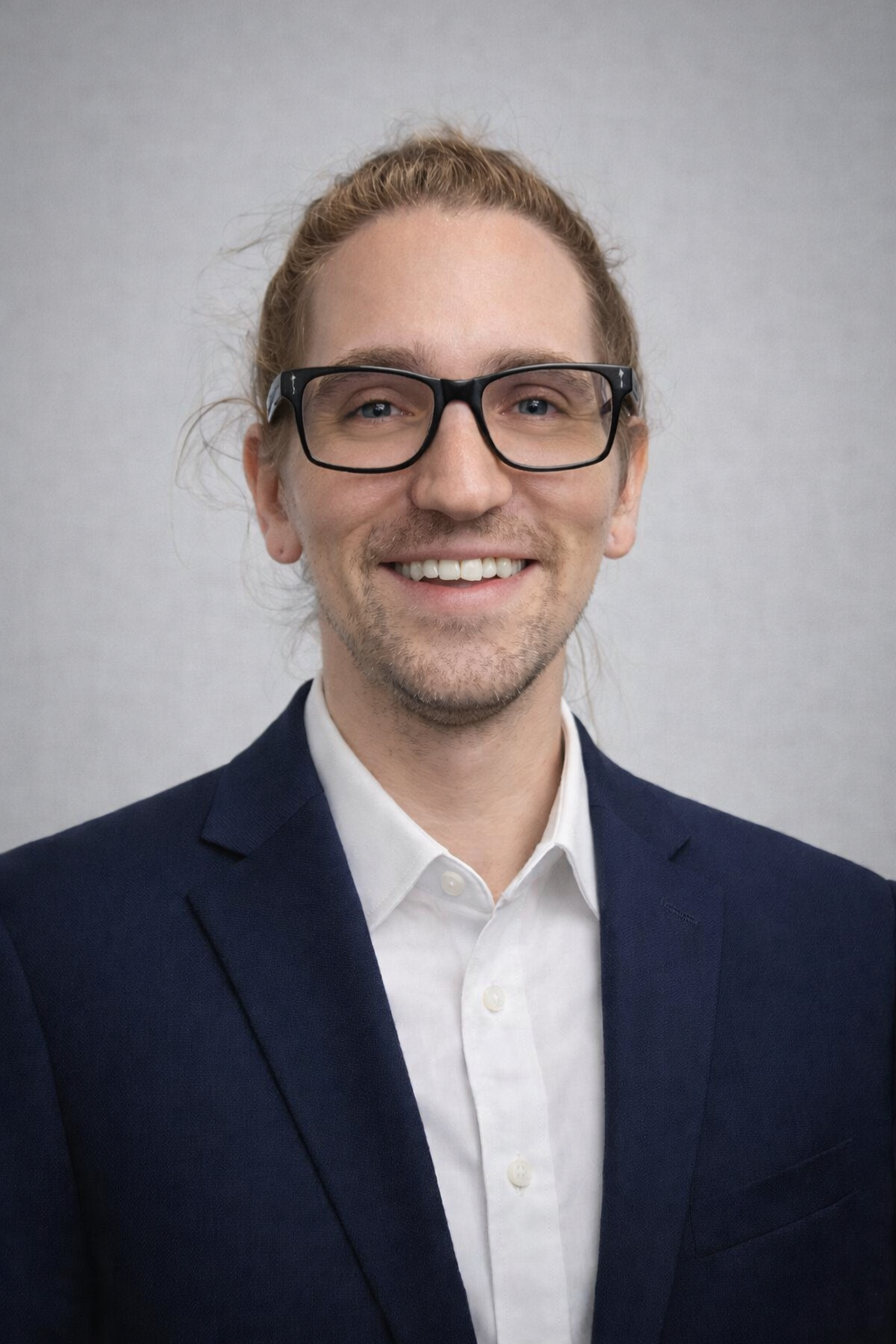 A person with long, light brown hair pulled back, wearing black glasses, a white shirt, and a navy blue blazer, smiles in front of a plain light gray background, representing Casillas Insurance Services in Katy TX.